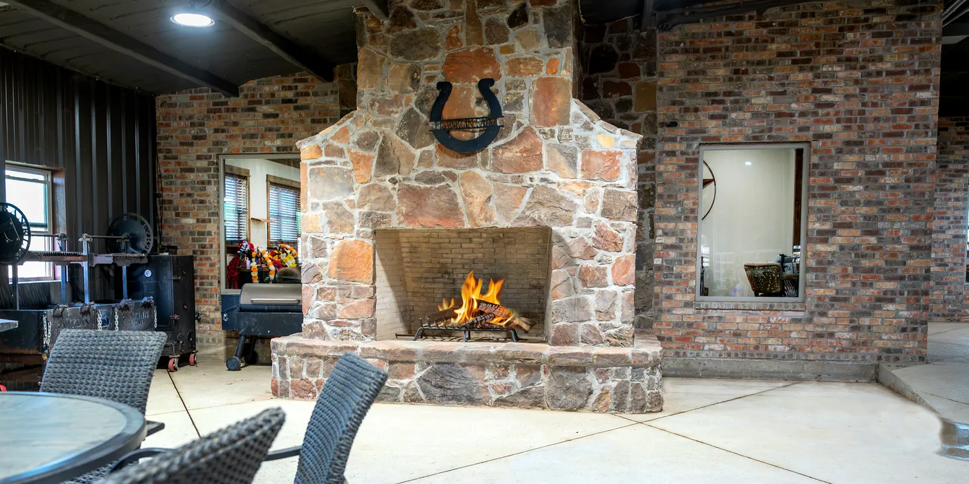 A covered lounge with a stone fireplace lit, a horseshoe above the hearth, and seating alongside.